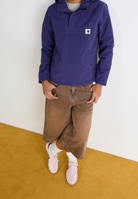 Persona con una chaqueta con capucha morada, pantalones marrones de pierna ancha hasta la rodilla, calcetines blancos y zapatillas rosas, de pie sobre un suelo amarillo.