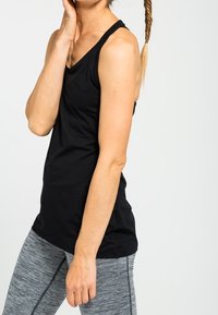 Černý bezrukávový top vyrobený z hladké tkaniny, s výstřihem do V. Nosený s šedými pruhovanými legínami. Modelka je v uvolněné póze.