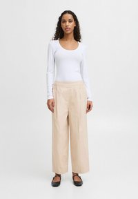 Top bianco a maniche lunghe e pantaloni beige a gamba larga. I pantaloni presentano una trama liscia e pieghe sottili. Ballerine nere con cinturini alla caviglia.