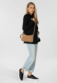 Braune Umhängetasche mit strukturierten Oberflächen, abgerundeten Ecken und einem dekorativen Quasten. Kombiniert mit einem schwarzen Blazer und hellblauen umgeschlagenen Jeans.