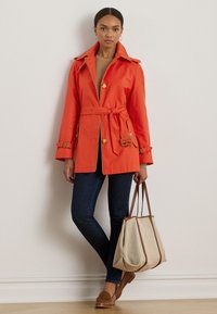 Feloranje, waterdichte trenchcoat met een riem, gouden accenten en knoopsluiting, gedragen met donkere jeans en een beige tote bag.