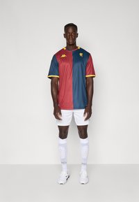 Sportstrøjen har et split-design i maroon og blå med gule accenter. Bæres med hvide shorts og sokker, hvilket viser en slank atletisk pasform.