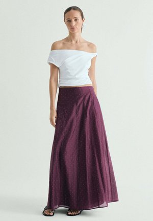 Mujer de pie con una blusa blanca sin hombros, falda larga morada con lunares y sandalias negras sobre un fondo liso claro.