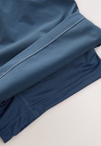 Atletski materijal tamnoplave boje s glatkom teksturom, sašivenim rubovima i slojevitim dizajnom. Unutrašnjost ima prozračnu mrežastu podstavu.