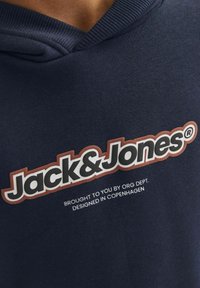 Tumesinine kapuutsiga särk, millel on silmapaistev valge ja punane "Jack & Jones" logo koos täiendava tekstiga all; kangas näeb välja sile ja pehme.