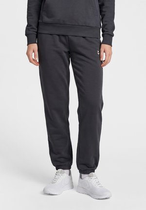 Person iført sorte joggingbukser med elastiske manchetter, kombineret med hvide sportssneakers og grå sokker på en ensfarvet baggrund.