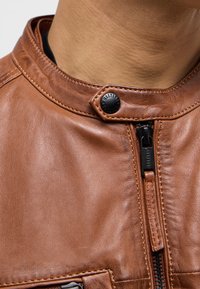 Braune Lederjacke mit hohem Kragen, Druckknopfverschluss und Vorderreißverschluss. Sichtbare Nähte und eine glatte, strukturierte Oberfläche.