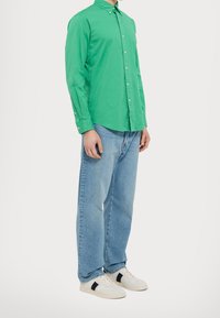 Zelená košile s dlouhým rukávem a knoflíky v kombinaci s světle modrými džíny a bílými teniskami s černými akcenty. Jednoduchý design a volný střih.