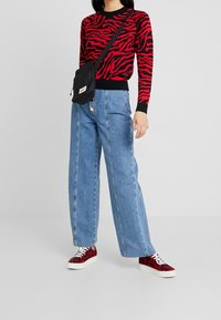 Svetr s černo-červeným zebrovým vzorem, světle modré široké kalhoty s vysokým pasem, černá crossbody kabelka a červené tenisky s leopardím potiskem.