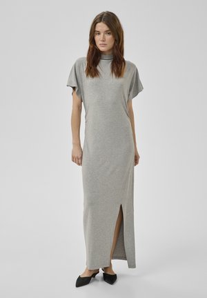 Femme debout portant une robe longue gris clair à manches courtes avec une fente latérale et des chaussures noires à bout pointu, sur un fond uni.