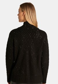 Zwarte gebreide trui met een hoge kraag, voorzien van een textuurpatroon van diagonale ribbels en open steken voor extra detail.