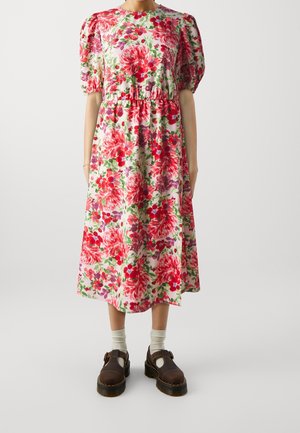 Kvinde iført en hvid kjole i mellemlængde med rød og lyserød blomsterprint, pufærmer, kombineret med hvide sokker og mørkebrune sko.