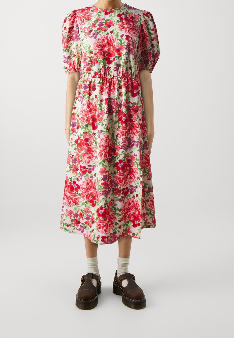 Femme portant une robe blanche mi-longue à imprimé floral rouge et rose, manches courtes bouffantes, associée à des chaussettes blanches et des chaussures marron foncé.
