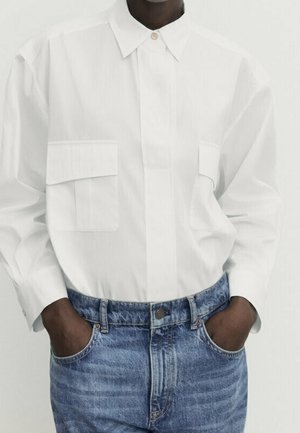 Person wearing a white button-up shirt with two front pockets tucked into high-waisted blue jeans with hands in pockets.