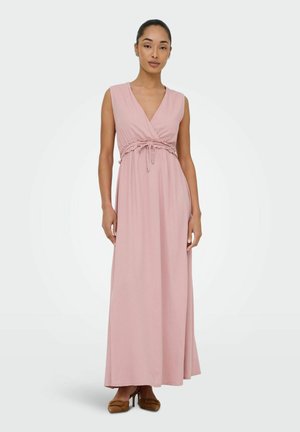 Žena stojaca v ružových dlhých šatách bez rukávov so sťahovacou šnúrkou v páse a hnedých lodičkách na vysokom opätku, na jednoduchom pozadí.