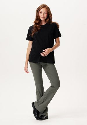 Jonge vrouw met lang bruin haar, gekleed in een zwart T-shirt, groene geribde broek en zwarte schoenen, staand met één hand op haar buik.