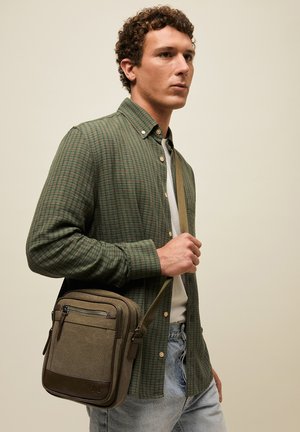 Noor mees, kes kannab rohelist ruudulist särki ja heleid teksaseid, kandes oliivirohelist õlakotti mitme taskuga.