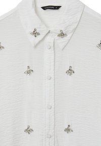 Camicia bianca con bottone, caratterizzata da una finitura texturizzata, con decorazioni in argento che ricordano api sul davanti.