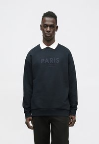 Sudadera azul marino con "PARIS" bordado en el pecho, cuello redondo y puños acanalados, combinada con una camisa blanca con cuello.