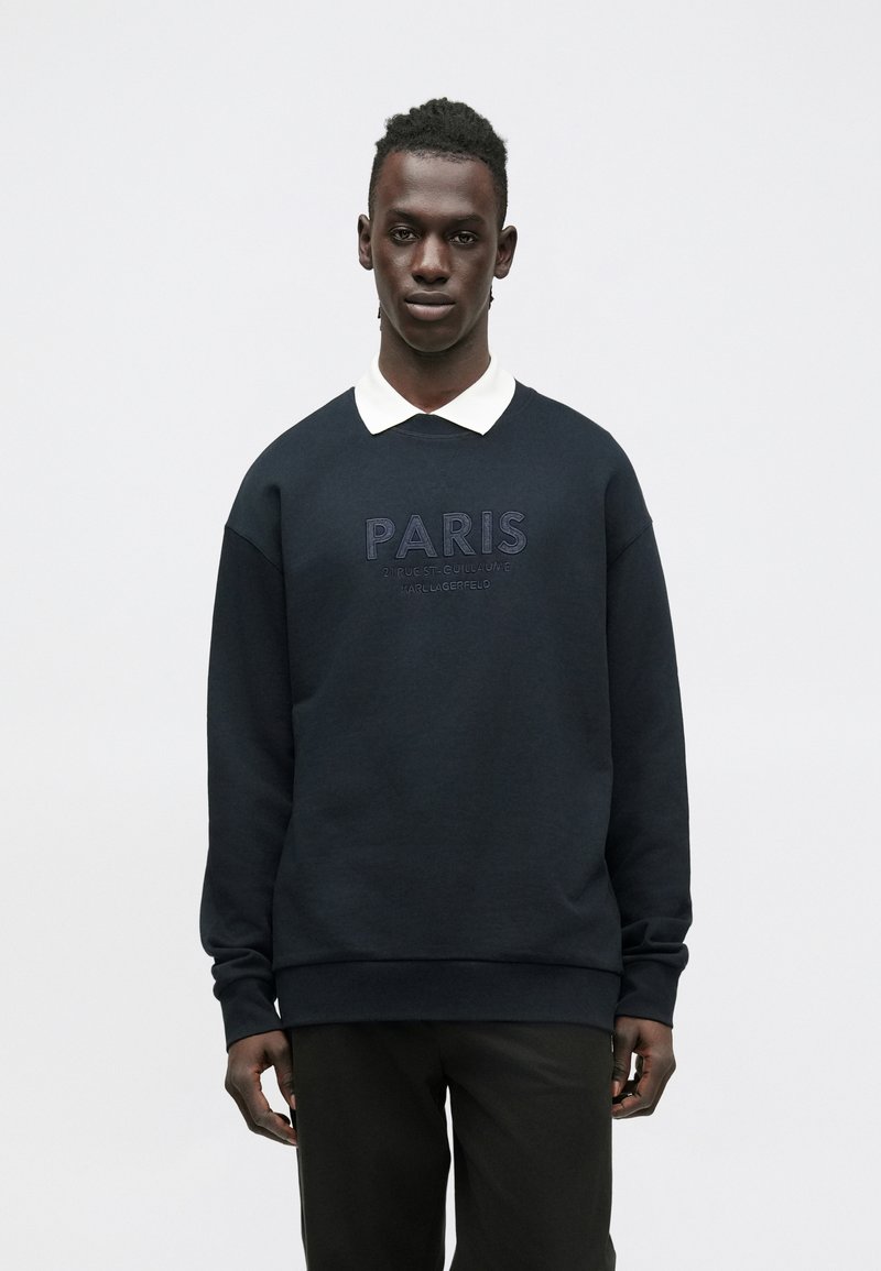 Sudadera azul marino con "PARIS" bordado en el pecho, cuello redondo y puños acanalados, combinada con una camisa blanca con cuello.