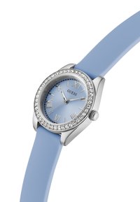 Relógio redondo prateado com mostrador azul claro, números romanos e correia de silicone azul. Destacado com cristais na luneta.