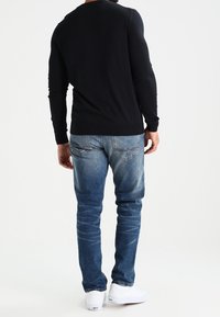 Mann in einem schwarzen Langarmshirt, blauen verwaschenen Jeans und weißen Sneakern, der mit dem Rücken zur Kamera steht.