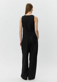 Femeie cu păr lung, castaniu, îmbrăcată cu un top fără mâneci cu dungi negre și pantaloni largi, stând cu mâinile în buzunare, întoarsă cu spatele.