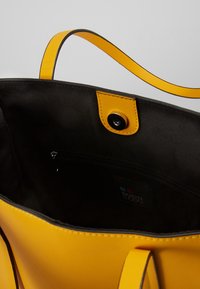 Borsa a spalla gialla con esterno liscio, fodera nera, tasca interna con cerniera e dettagli a strappo gialli a contrasto. Chiusura a scatto magnetico.