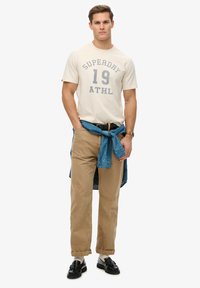 Camiseta beige con letras grises, pantalones caqui y una camisa de mezclilla azul atada a la cintura. Zapatillas negras completan el atuendo.