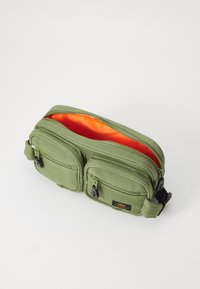 Λαδί υφασμάτινη τσάντα μέσης με δύο φερμουάρ, πορτοκαλί εσωτερικό και μαύρα μεταλλικά στοιχεία. Συμπεριλαμβάνεται ρυθμιζόμενος ιμάντας.