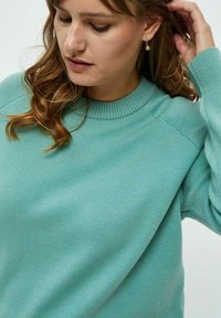 Maglione turchese a maglia con scollatura rotonda, polsini a coste e tessuto testurizzato, abbinato a orecchini di perle pendenti.