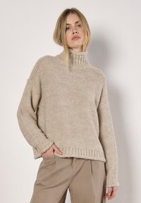Beige neulepusero, jossa on korkea, ribbikuvioitu kaulus, pudotetut olkapäät ja leveät hihat. Raskaasti teksturoitu, löysä malli, jossa ribbikuvioitu helma ja hihansuut.