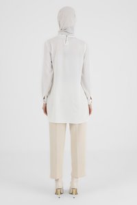 White long tunic with a high neckline and back button closure, paired with beige trousers. Textured fabric with loose sleeves and cuffs.