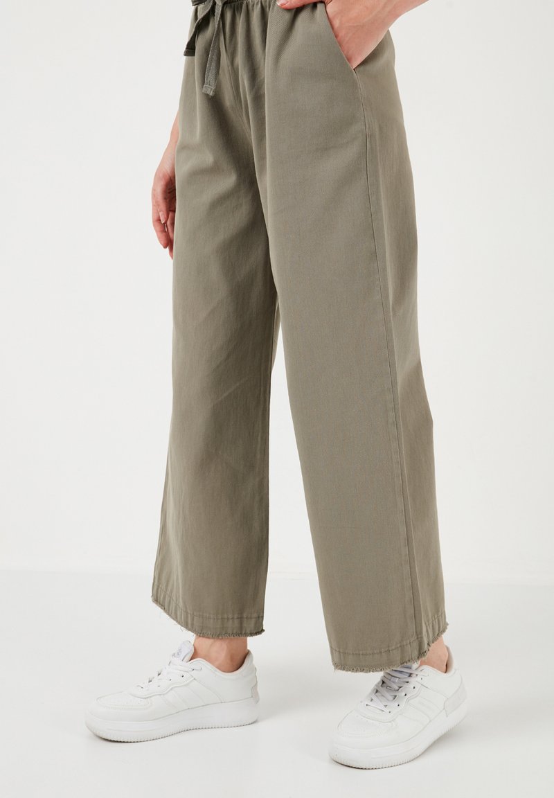 Licht olijfgroene, wijde broek met een trekkoord in de taille, voorzien van zijzakken en rauwe zomen, gecombineerd met witte sneakers.
