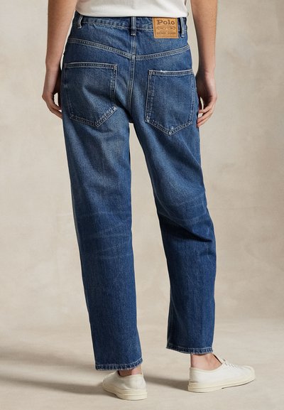 Persona con vaqueros azules de pierna recta, bolsillos traseros y una etiqueta de cuero Polo Denim Goods, combinados con zapatillas blancas y una camisa blanca.