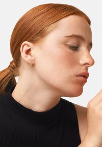 Goldene Creole an einem Model. Die Ohrringe haben ein minimalistisch geometrisches Design. Das Model hat helle Haut, rote Haare und trägt ein dunkles geripptes Oberteil.