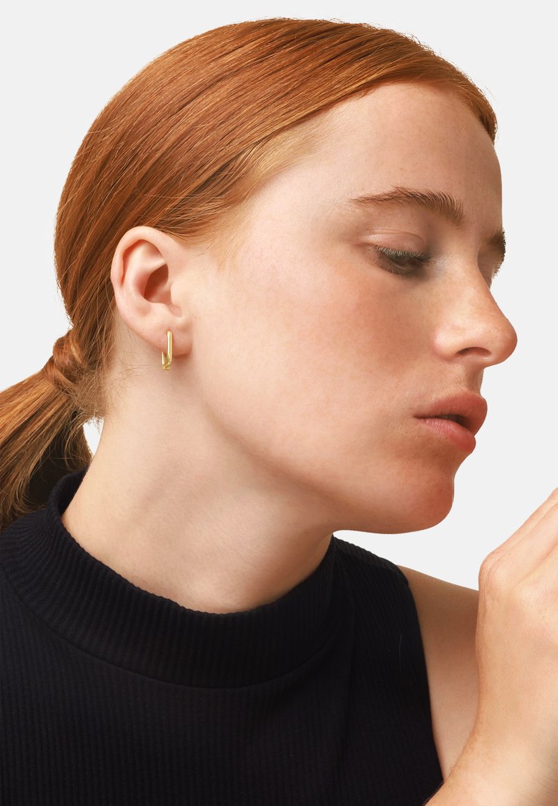 Goldene Creole an einem Model. Die Ohrringe haben ein minimalistisch geometrisches Design. Das Model hat helle Haut, rote Haare und trägt ein dunkles geripptes Oberteil.