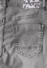 Los jeans de mezclilla gris presentan un bolsillo trasero estándar con un borde cosido y un pequeño remache metálico. La tela tiene una textura suave y ligeramente deslavada.