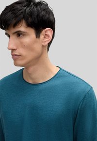Türkisfarbenes Langarmshirt mit rundem Ausschnitt, aus weichem Material, mit subtiler Textur und minimalen Nähdetails.