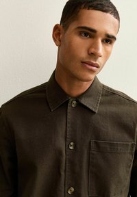 Camisa de algodón marrón con cuello puntiagudo, botones frontales y bolsillo en el pecho. Textura suave con sutiles variaciones tonales.