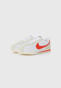 Sportiniai bateliai su balta viršutine dalimi, raudonu swoosh logotipu ir grietinėlės spalvos zomšiniais akcentais. Padas yra šviesiai rudos spalvos su tekstūruotu raštu.