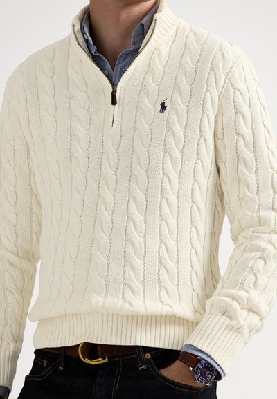 Elfenbensfarget kabelstrikket genser med kvart-Zip-krage, ribbede mansjetter og kant, med en liten logo på brystet. Brukes over en lyseblå skjorte.