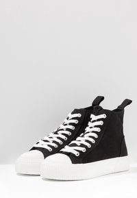 Sorte højtop sneakers lavet af lærred med hvide snørebånd, gummitåhætte og tyk hvid sål. Har trækflapper på hælen.