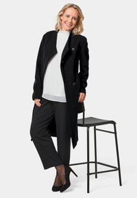 Schwarzer Wollmantel mit drapierter Front und zwei Knöpfen, kombiniert mit einer weißen Bluse und schwarzen, maßgeschneiderten Hosen. Schwarze High Heels runden das Outfit ab.