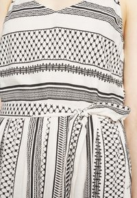 Robe à motifs en noir et blanc comportant un décolleté en V, une taille cintrée avec une ceinture nouée, et un tissu texturé avec des motifs géométriques.