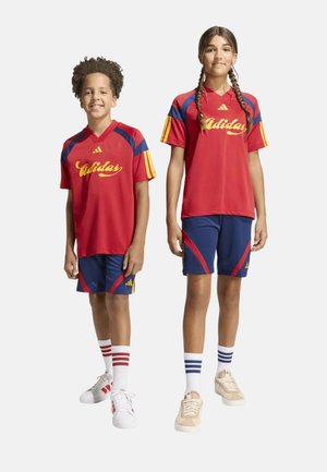 Dos niños sonrientes que llevan camisetas deportivas rojas a juego de Adidas con logo amarillo, pantalones cortos azules, calcetines blancos y zapatillas, de pie uno al lado del otro.