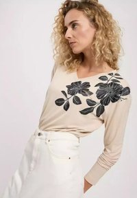 Beżowa bluzka z długim rękawem i dekoltem w kształcie V z czarnym haftem kwiatowym na jednym ramieniu; zestawiona z białą spódnicą o wysokim stanie.