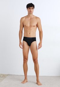 Schwarze Baumwollslips mit einem mit Logo verzierten Bund, glattem Stoff und einer figurbetonten Passform, präsentiert an einem männlichen Model.