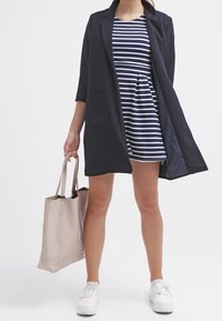 Laivastonsininen pitkähihainen bleiseri avonaisella etuosalla, yhdistettynä raidalliseen mekkoon. Mallilla on käsissään vaaleanruskea olkalaukku ja jalassaan valkoiset tennarit. Pintarakennus on sileä.