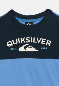 Μπλουζάκι σε μπλε ναυτικό και ανοιχτό μπλε χρώμα με το λογότυπο Quiksilver, γραφικό κύμα και το κείμενο "ESTD. 1969" στο στήθος.
